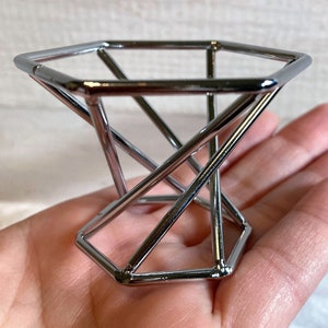 May include: A silver metal makeup sponge holder with a geometric design. The holder is shaped like a pyramid with a hexagonal base.