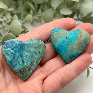 May include: Two heart-shaped stones with a turquoise and green color scheme. The stones are held in a hand, showcasing their unique patterns and textures. The heart-shaped stones are a decorative item.