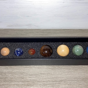 May include: A set of eight small, round, polished stones in a black box with a black foam interior. The stones are various colors, including white, orange, blue, red, brown, yellow, green, and light blue.