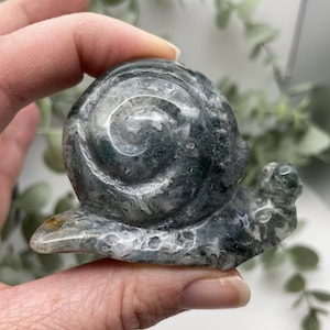 May include: A green and gray agate snail sculpture. The snail is carved from a single piece of agate and has a detailed shell and body.
