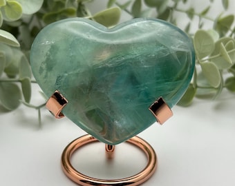 May include: A heart-shaped, translucent fluorite stone in shades of green and blue, mounted on a copper-colored metal stand. The stone has natural veining and is displayed against a backdrop of green foliage.