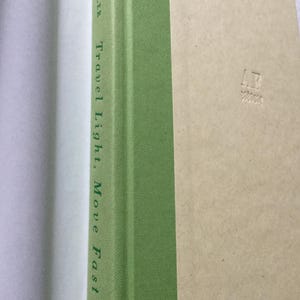 Puede incluir: Un libro titulado "Travel Light, Move Fast" de Laura Fuller. El lomo es verde con texto blanco. La portada es de color beige claro con las iniciales "AR" en relieve en la esquina inferior derecha. El libro est&aacute; abierto, revelando p&aacute;ginas blancas.