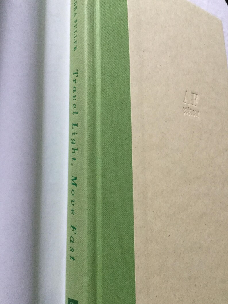 Puede incluir: Un libro titulado "Travel Light, Move Fast" de Laura Fuller. El lomo es verde con texto blanco. La portada es de color beige claro con las iniciales "AR" en relieve en la esquina inferior derecha. El libro est&aacute; abierto, revelando p&aacute;ginas blancas.
