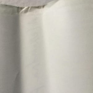 Puede incluir: Primer plano de un papel blanco doblado. El papel tiene una ligera textura y est&aacute; arrugado en varios lugares. El borde superior del papel es visible, mostrando una costura donde est&aacute; unido a otro material.
