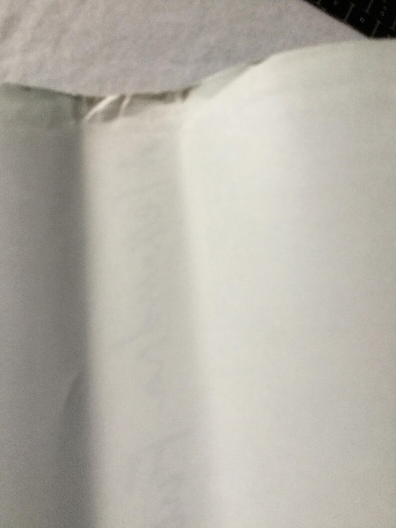 Puede incluir: Primer plano de un papel blanco doblado. El papel tiene una ligera textura y est&aacute; arrugado en varios lugares. El borde superior del papel es visible, mostrando una costura donde est&aacute; unido a otro material.