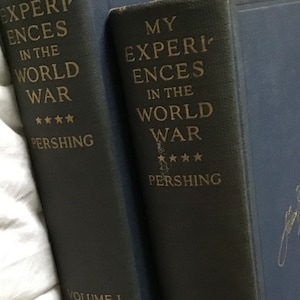 May include: Two dark blue hardcover books with gold lettering. The books are titled "My Experiences in the World War" by Pershing. The first book is labeled "Volume I" and the second book is labeled "Volume II".