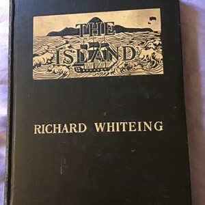 Op de afbeelding: Een zwart hardback boek met gouden letters. De titel is "THE ISLAND" met een zwart-witte illustratie van een schip dat op zee vaart. De auteur is "RICHARD WHITEING".