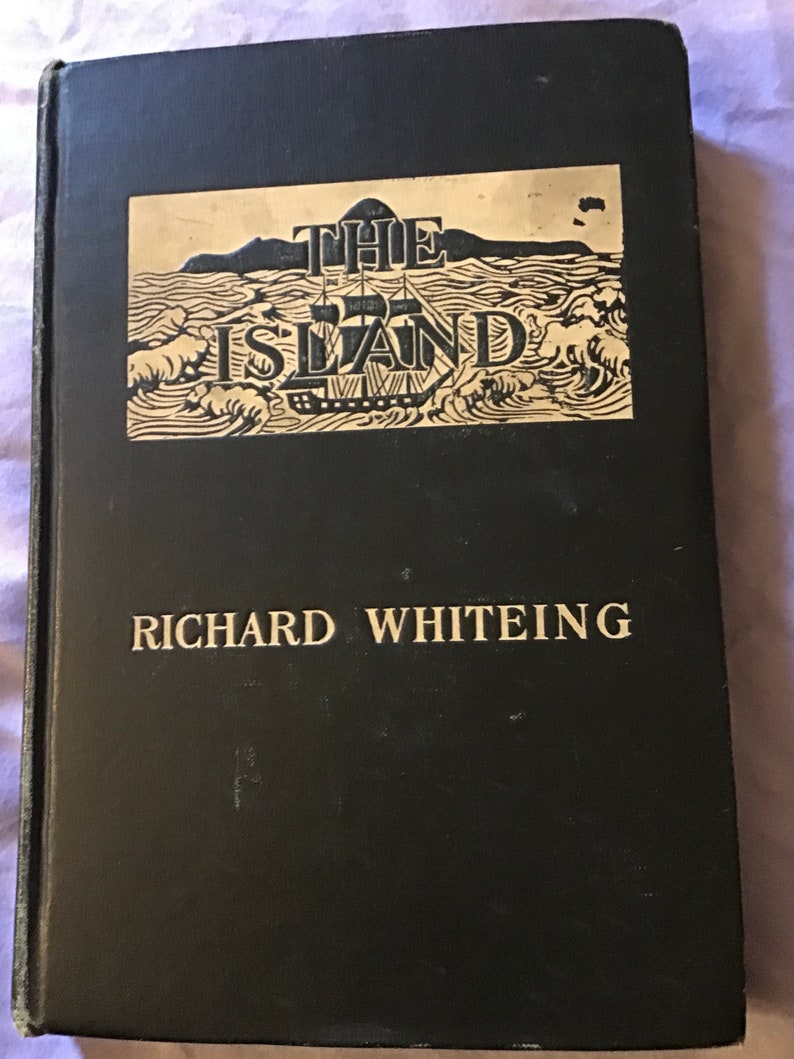 Op de afbeelding: Een zwart hardback boek met gouden letters. De titel is "THE ISLAND" met een zwart-witte illustratie van een schip dat op zee vaart. De auteur is "RICHARD WHITEING".