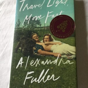 Puede incluir: Portada del libro "Travel Light, Move Fast" de Alexandra Fuller. La imagen muestra una pareja y un perro sobre hierba, con el t&iacute;tulo en escritura blanca. Una pegatina de librer&iacute;a es visible. El fondo es verde y azul.