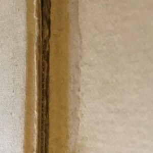 Op de afbeelding: Close-up van een naad tussen twee stukken karton. De naad is gemaakt van een dunne strook karton die aan de randen van de twee stukken is gelijmd. Het karton is lichtbruin.