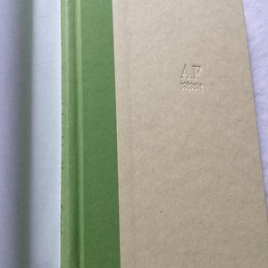 Puede incluir: Un libro de tapa dura con lomo verde claro y cubierta beige. La cubierta presenta las iniciales "A.F." en relieve y un patr&oacute;n en zigzag debajo. El lomo del libro es de color verde claro liso.
