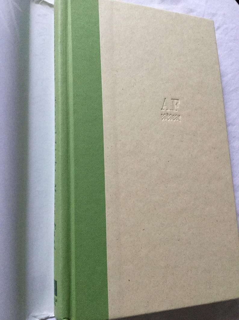 Puede incluir: Un libro de tapa dura con lomo verde claro y cubierta beige. La cubierta presenta las iniciales "A.F." en relieve y un patr&oacute;n en zigzag debajo. El lomo del libro es de color verde claro liso.