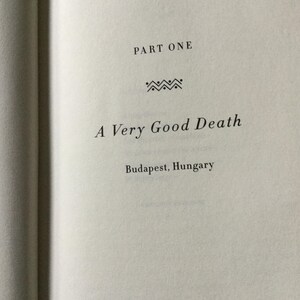 Puede incluir: P&aacute;gina de libro abierta con el t&iacute;tulo "A Very Good Death" y la ubicaci&oacute;n "Budapest, Hungr&iacute;a". El texto est&aacute; en una fuente serif, con "PART ONE" en la parte superior. Una l&iacute;nea decorativa est&aacute; entre el t&iacute;tulo y "PART ONE".