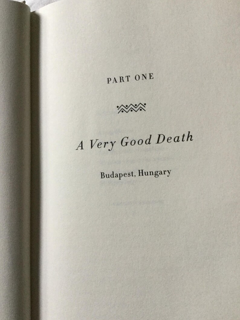 Puede incluir: P&aacute;gina de libro abierta con el t&iacute;tulo "A Very Good Death" y la ubicaci&oacute;n "Budapest, Hungr&iacute;a". El texto est&aacute; en una fuente serif, con "PART ONE" en la parte superior. Una l&iacute;nea decorativa est&aacute; entre el t&iacute;tulo y "PART ONE".