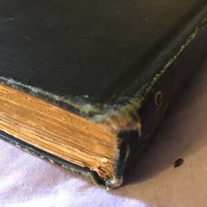Op de afbeelding: Close-up van de rug van een zwart hardback boek met goudkleurige pagina's. Het boek is licht beschadigd en heeft een paar kleine scheuren op de rug.