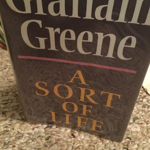 May include: A book cover with the title "A Sort of Life" by Graham Greene. The title is in gold lettering on a dark brown background.