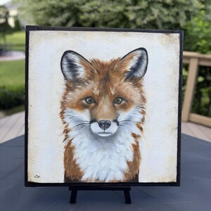 May include: A square painting of a fox head and chest on a light cream background, framed in a thin black border. The fox features reddish-brown fur, white chest markings, pointed ears, and dark eyes. The artist's initials are visible in the bottom left.