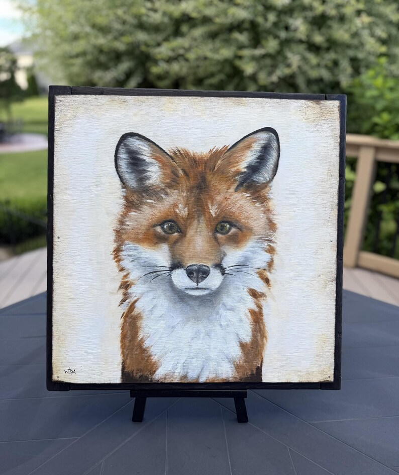 May include: A square painting of a fox head and chest on a light cream background, framed in a thin black border. The fox features reddish-brown fur, white chest markings, pointed ears, and dark eyes. The artist's initials are visible in the bottom left.