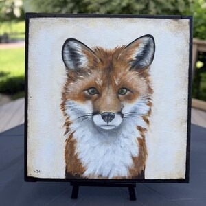 May include: A square painting of a fox head and chest on a light cream background, framed in a thin black border. The fox features reddish-brown fur, white chest markings, pointed ears, and dark eyes. The artist's initials are visible in the corner.