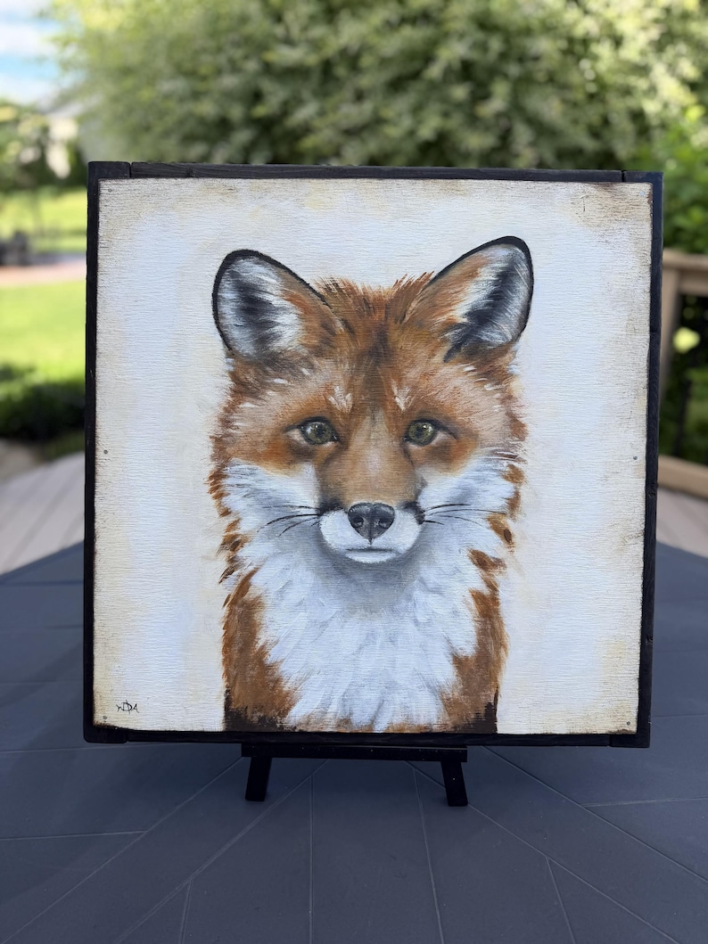 May include: A square painting of a fox head and chest on a light cream background, framed in a thin black border. The fox features reddish-brown fur, white chest markings, pointed ears, and dark eyes. The artist's initials are visible in the corner.
