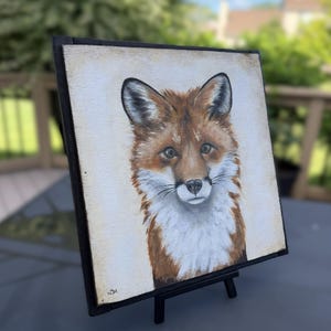 May include: A square painting of a fox head and chest featuring reddish-brown fur, white chest markings, and dark-tipped ears. The artwork is set within a black wooden frame and is displayed on a small black easel, showing a signature in the corner.