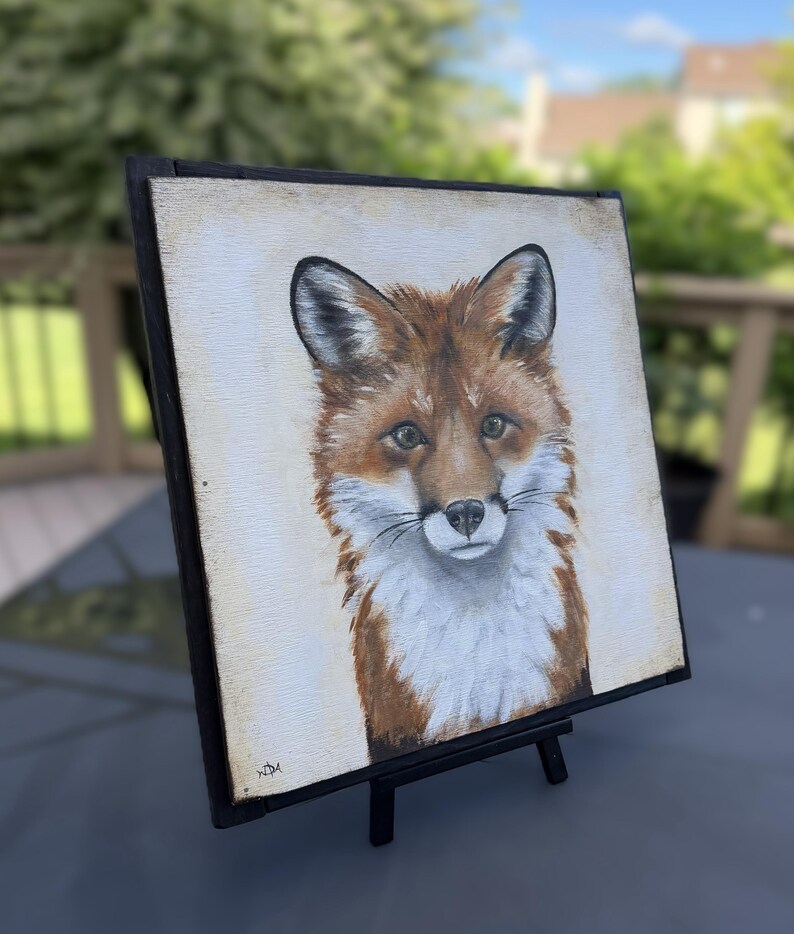 May include: A square painting of a fox head and chest featuring reddish-brown fur, white chest markings, and dark-tipped ears. The artwork is set within a black wooden frame and is displayed on a small black easel, showing a signature in the corner.