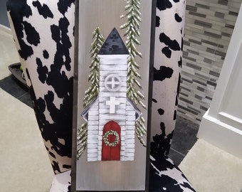 May include: A hand-painted wooden sign with a white church building and two green pine trees with snow on them. The church has a red door with a green wreath and a white cross on the front. The sign has a distressed gray background.