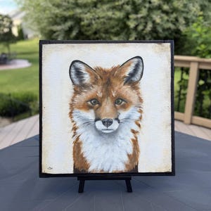 May include: A square painting of a fox head and chest featuring reddish-brown fur, white chest markings, and dark-tipped ears. The fox is centered on a light cream background within a thin black frame, resting on a small black display easel.