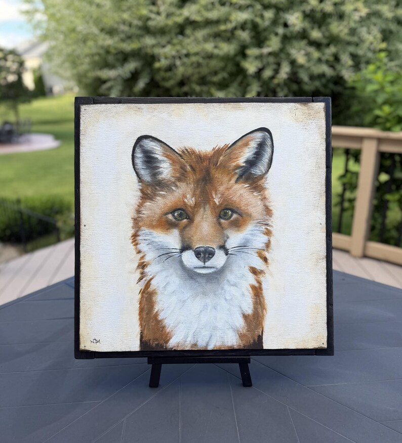 May include: A square painting of a fox head and chest featuring reddish-brown fur, white chest markings, and dark-tipped ears. The fox is centered on a light cream background within a thin black frame, resting on a small black display easel.