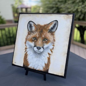 May include: A square painting on a black frame featuring a detailed portrait of a red fox with white fur on its chest and face, dark-tipped ears, and brown eyes, set against a light cream-colored background with a small signature in the corner.