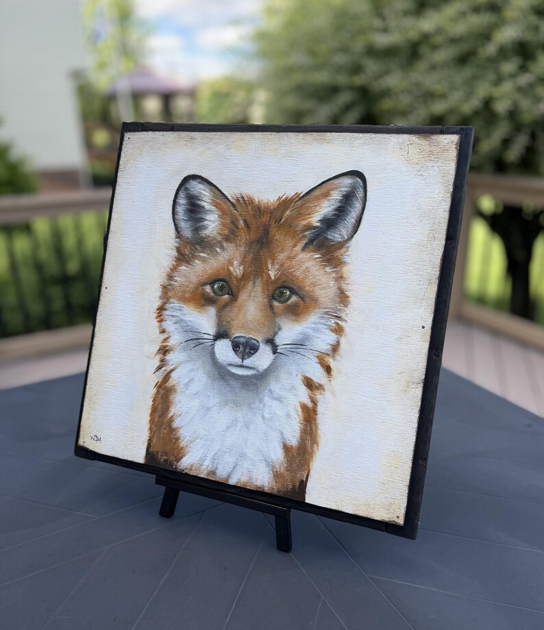 May include: A square painting on a black frame featuring a detailed portrait of a red fox with white fur on its chest and face, dark-tipped ears, and brown eyes, set against a light cream-colored background with a small signature in the corner.