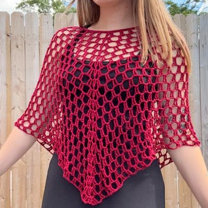 Puede incluir: Un poncho de ganchillo rojo oscuro con un diseño de red. El poncho es de color rojo intenso y tiene forma cuadrada. La prenda se lleva sobre una camiseta negra.