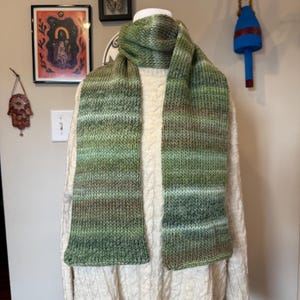 May include: A green and brown knitted scarf displayed on a mannequin. The scarf has a striped pattern with shades of green, brown, and white. The scarf is made of a soft, textured yarn, perfect for cold weather.
