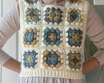 Può includere: Un gilet all'uncinetto con un motivo a quadrati della nonna in tonalità di bianco, verde e giallo. Il gilet ha una scollatura rotonda ed è senza maniche.