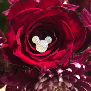 Pode incluir: Um close-up de um buquê com uma rosa vermelha escura com uma cabeça de Mickey Mouse brilhante coberta de strass no centro. A rosa é cercada por crisântemos roxos escuros, criando um contraste vibrante.