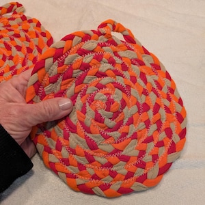 May include: Two round, braided pot holders in orange, red, and beige. The pot holders are made of fabric and have a woven design.