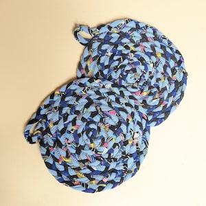 May include: Two round, braided fabric trivets featuring a mix of light blue, black, and multi-colored patterned textile strips. Each trivet is constructed from coiled, stitched fabric and includes a small fabric loop for hanging storage.
