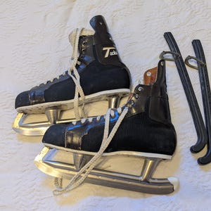 May include: A pair of vintage black ice skates with silver blades and white laces. The skates have a high-top design with the word "Tacks" printed on the side. Two black skate guards are also included.