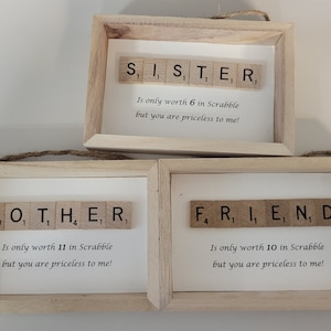 Puede incluir: Tres marcos de madera con fondos blancos y fichas de Scrabble que deletrean "Hermana", "Madre" y "Amigo". Cada marco incluye un mensaje sobre la puntuación de Scrabble de la palabra y su valor incalculable.