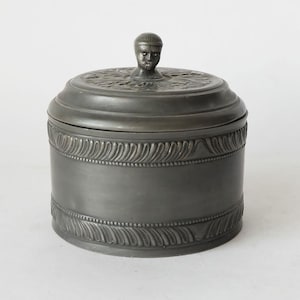 Op de afbeelding: Een tinnen pot met een deksel met een gebeeldhouwd mensenhoofd. De pot heeft een decoratief ontwerp rond de boven- en onderkant.