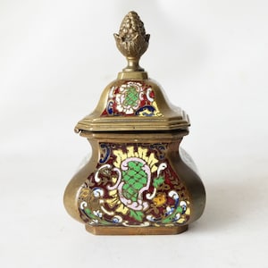 Könnte beinhalten: Eine kleine, quadratische, verzierte Dose aus Messing mit Deckel und einem dekorativen Knauf. Die Dose ist mit bunten Emaille-Blumenmustern in Rot-, Grün-, Gelb- und Blautönen verziert.