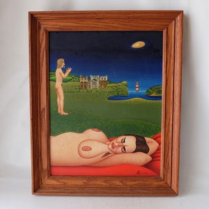 Op de afbeelding: Een ingelijst schilderij dat een surrealistisch landschap afbeeldt met een vrouw die op een rode deken ligt, een man die in de verte staat en een kasteel op de achtergrond. Het schilderij is geplaatst tegen een donkerblauwe lucht met een halve maan.