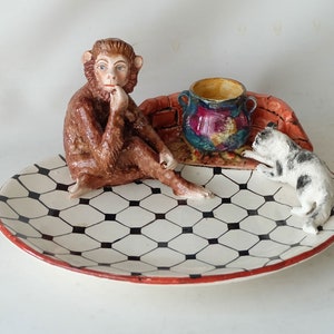 以下が含まれることがあります： 黒と白の市松模様のセラミックプレート。プレートの上に座っている茶色の猿の置物、カラフルなデザインの小さなセラミックポット、黒と白の猫の置物が置かれている。