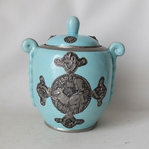 Peut inclure: Un pot en céramique bleu clair avec un couvercle et deux anses. Le pot est décoré d'accents en métal argenté dans un motif de nœud celtique. Le pot porte une inscription en relief qui dit "CAPEL".