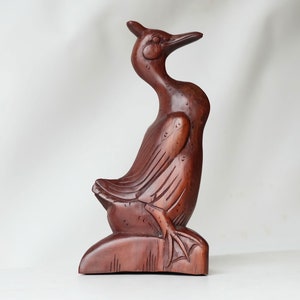 Op de afbeelding: Een handgesneden houten vogelsculptuur in donkerbruine kleur. De vogel zit op een voetstuk en heeft zijn kop omhoog gekanteld.