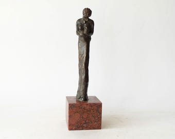 May include: A bronze sculpture of a person standing on a red marble base. The sculpture is abstract and depicts a person with their arms crossed.