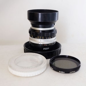 May include: A vintage camera lens with a black and silver body, a black lens hood, and a 52mm Nikon filter. A clear glass lens protector sits beside the lens. The lens has various measurement markings.