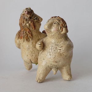 May include: A ceramic sculpture of two figures with human-like features. The figures are beige with brown and reddish-brown accents, and appear to be embracing. The sculpture has a textured surface and is set against a plain background.