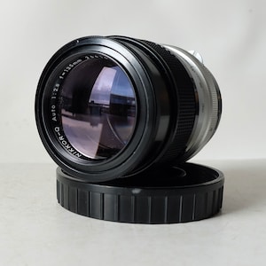 May include: A black Nikon-Q Auto 1:2.8 f=135mm lens with a silver band around the middle. The lens is resting on a black lens cap.