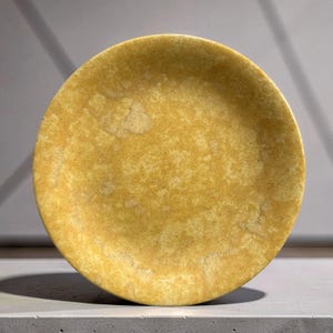 Op de afbeelding: Een rond bord met een gespikkeld mosterdgeel oppervlak met een getextureerde, onregelmatige afwerking. Het bord heeft een licht opstaande rand en een consistent kleurpatroon over het gehele oppervlak, met variaties in lichte en donkere geeltinten.